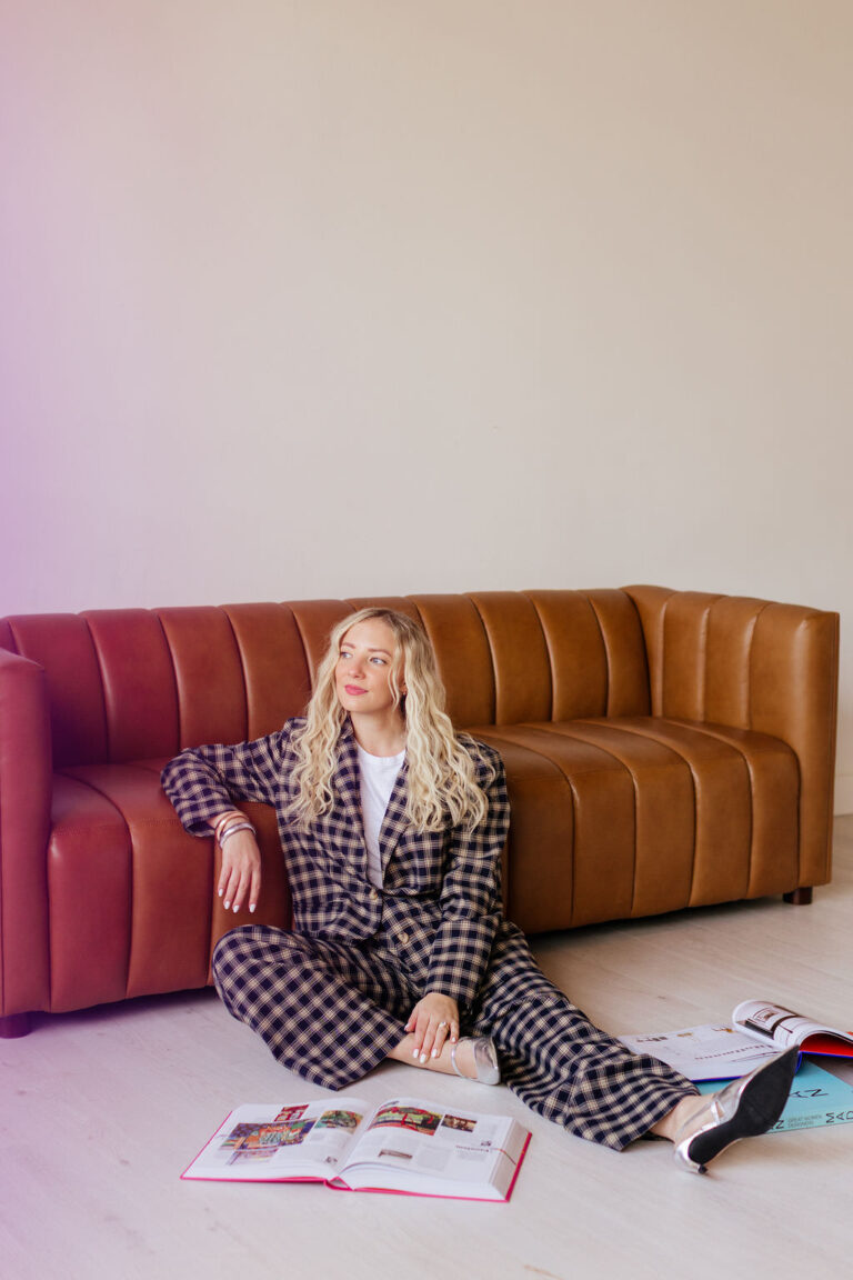 A woman sits on the floor leaning on a leather couch with books on the floor around her