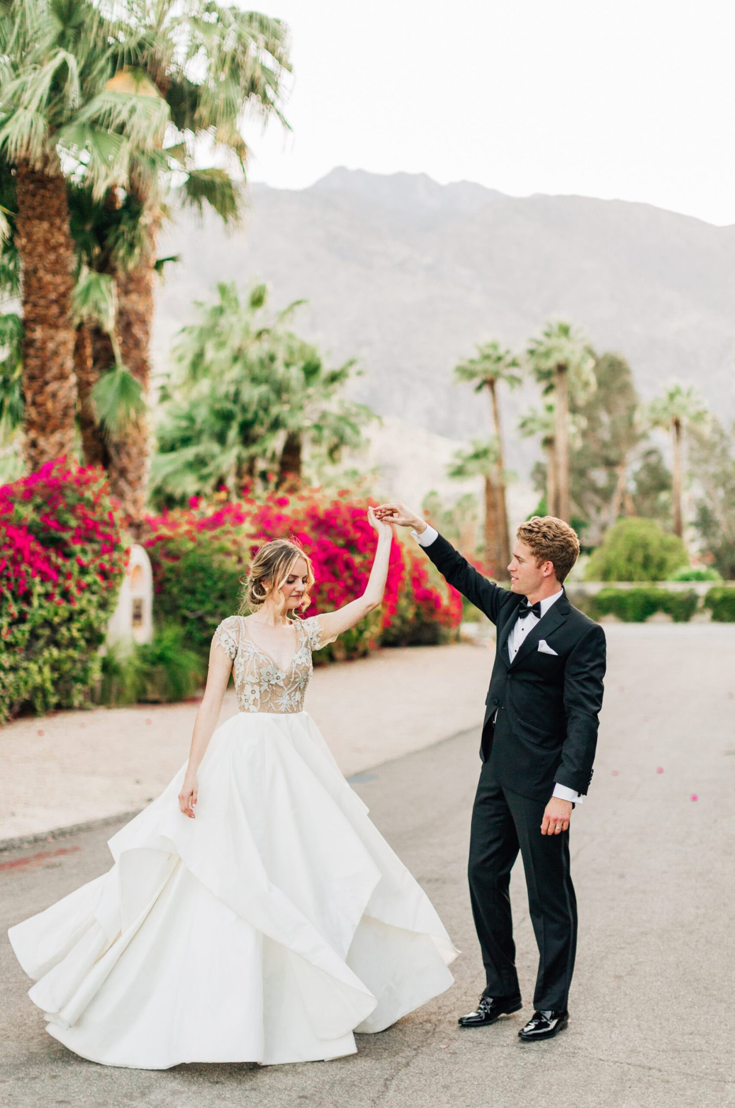 A bride and groom twirl and dance in the street with flowers and palm springs mountains in the distance