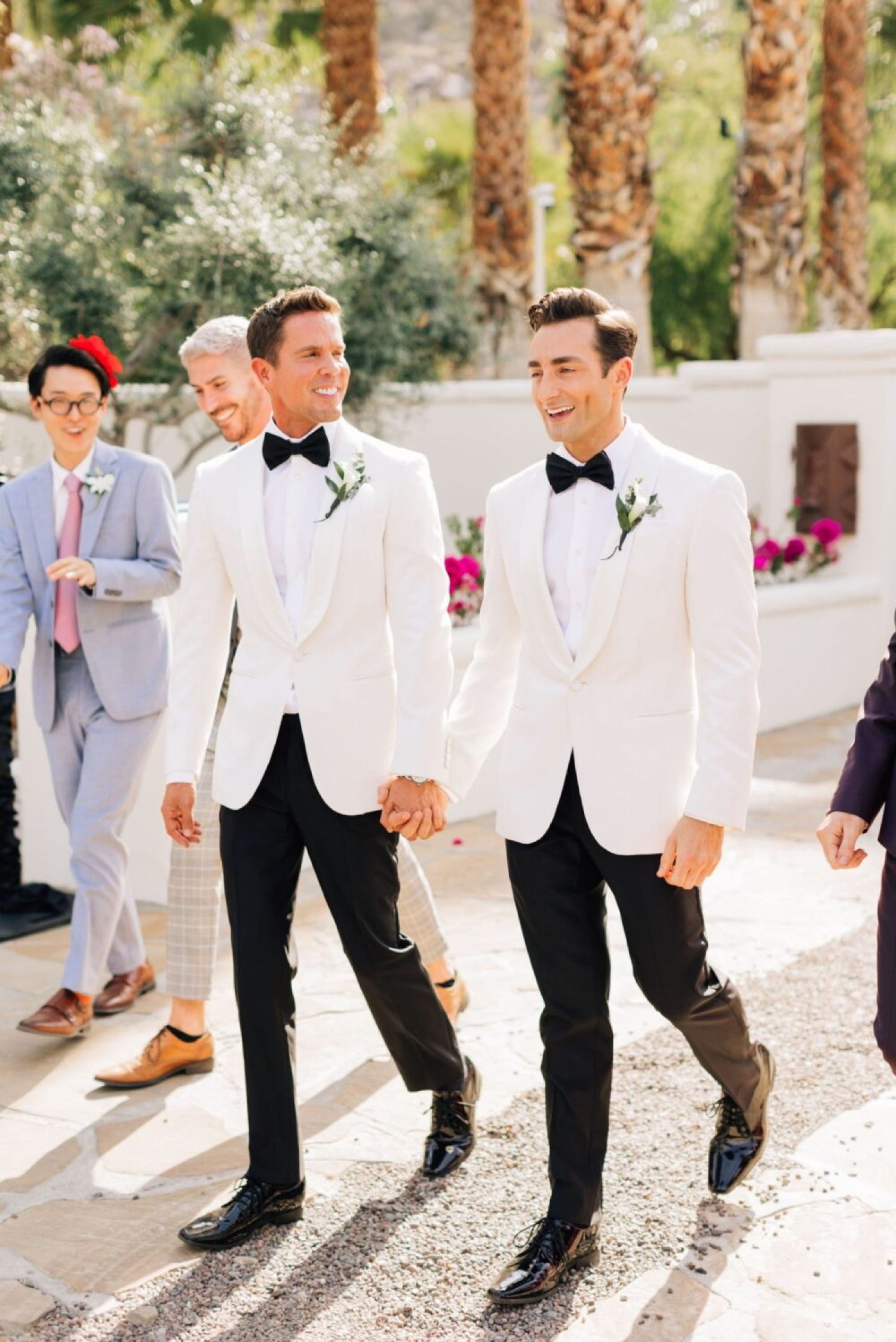 Two grooms and their wedding party walk down a path in white tuxedos in palm springs shot by a los angeles wedding photographer
